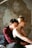 A man and a woman are sitting on a bed, with the man embracing the woman from behind. The man is wearing a maroon long-sleeve shirt and has short dark hair, whereas the woman has light brown hair and is wearing a black sleeveless top. The setting seems to be a rustic or dilapidated room with peeling paint on the walls and a light fixture hanging above them.