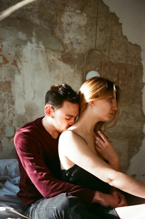 A man and a woman are sitting on a bed, with the man embracing the woman from behind. The man is wearing a maroon long-sleeve shirt and has short dark hair, whereas the woman has light brown hair and is wearing a black sleeveless top. The setting seems to be a rustic or dilapidated room with peeling paint on the walls and a light fixture hanging above them.