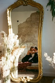 A large vintage mirror with an ornate golden frame reflects a couple sitting closely together, suggesting affection or intimacy. They are surrounded by warmly lit, cozy decor including tall pampas grass and an exposed, textured wall in the background.