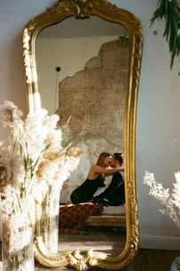 A large vintage mirror with an ornate golden frame reflects a couple sitting closely together, suggesting affection or intimacy. They are surrounded by warmly lit, cozy decor including tall pampas grass and an exposed, textured wall in the background.