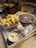 A glass of amber beer sits on a wooden tray decorated with newspaper print. Next to the beer is a container filled with golden French fries and a small bowl of creamy sauce. The setting is a rustic, wooden café with visible wooden chairs and a warm ambiance.
