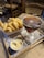 A glass of amber beer sits on a wooden tray decorated with newspaper print. Next to the beer is a container filled with golden French fries and a small bowl of creamy sauce. The setting is a rustic, wooden café with visible wooden chairs and a warm ambiance.