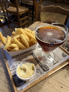 A glass of amber beer sits on a wooden tray decorated with newspaper print. Next to the beer is a container filled with golden French fries and a small bowl of creamy sauce. The setting is a rustic, wooden café with visible wooden chairs and a warm ambiance.