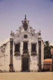 A historic white stucco church with ornate architectural details, including decorative stonework around windows and a rounded roofline. The facade shows signs of weathering and age, with a palm tree visible behind the structure. The sky is clear and blue, and a cobblestone area lies in front, contributing to an atmosphere of antiquity.