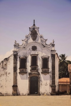 A historic white stucco church with ornate architectural details, including decorative stonework around windows and a rounded roofline. The facade shows signs of weathering and age, with a palm tree visible behind the structure. The sky is clear and blue, and a cobblestone area lies in front, contributing to an atmosphere of antiquity.