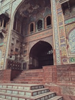 A detailed architectural facade featuring intricate tile work and ornate patterns. The building includes a large archway entry with decorative elements and a series of steps leading up to it. The walls are adorned with colorful floral and geometric designs, with a blend of earth tones and vivid colors.