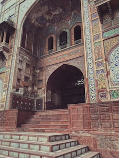 A detailed architectural facade featuring intricate tile work and ornate patterns. The building includes a large archway entry with decorative elements and a series of steps leading up to it. The walls are adorned with colorful floral and geometric designs, with a blend of earth tones and vivid colors.