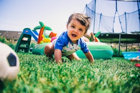 A baby in a blue shirt is joyfully crawling on green grass in a backyard, with a colorful inflatable pool, small slide, trampoline, and a soccer ball nearby, creating a playful and lively atmosphere.