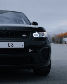 A black Land Rover SUV is parked on a street with its headlights on. The front view of the vehicle reveals details such as the grille and logo. The background shows a road that extends towards a mountainous landscape with trees on the sides, highlighted by overcast weather.