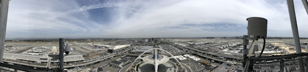 An expansive aerial view of an airport, showcasing multiple runways, terminals, and aircraft parked at gates. The scene is bustling with various vehicles and infrastructure including roads weaving between the buildings. The architecture features a prominent, distinctive, futuristic building with curves at the center, surrounded by a vast network of air traffic areas. The sky above is partly cloudy with a few streaks of cirrus clouds.