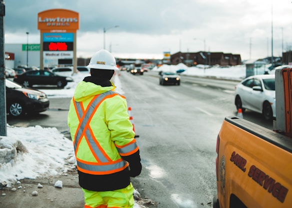 A person in a bright neon safety jacket and hard hat stands on a snowy sidewalk, watching over a road where cars are passing by. Nearby, a service vehicle is parked, and there's a large retail sign for Lawtons Drugs visible in the background. The scene is set in a wintry environment with snow visible on the ground.