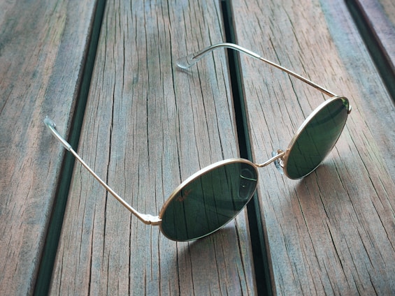 A pair of round sunglasses with gold frames and dark lenses rests on a rustic wooden surface. The wood planks have visible grain and a weathered appearance, adding texture to the background.