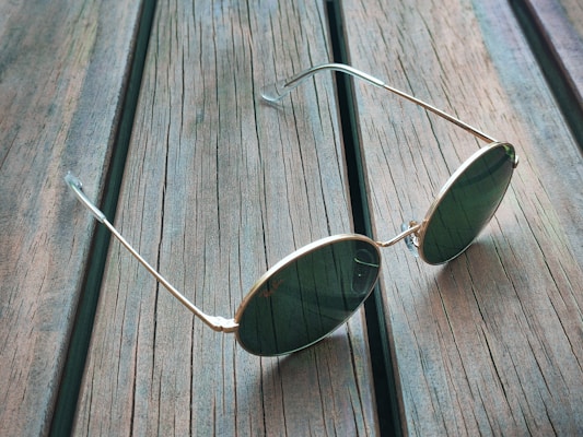 A pair of round sunglasses with gold frames and dark lenses rests on a rustic wooden surface. The wood planks have visible grain and a weathered appearance, adding texture to the background.