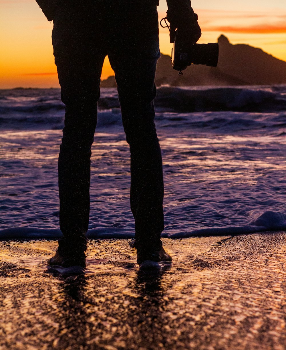 silhueta da pessoa segurando a câmera em pé na costa durante o pôr do sol