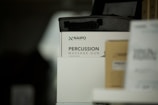 A product display featuring a percussion massage gun in its packaging. The box is predominantly white with the brand and product name clearly visible. There are other boxes partially visible around it, one of which is brown.