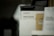 A product display featuring a percussion massage gun in its packaging. The box is predominantly white with the brand and product name clearly visible. There are other boxes partially visible around it, one of which is brown.