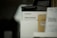 A product display featuring a percussion massage gun in its packaging. The box is predominantly white with the brand and product name clearly visible. There are other boxes partially visible around it, one of which is brown.