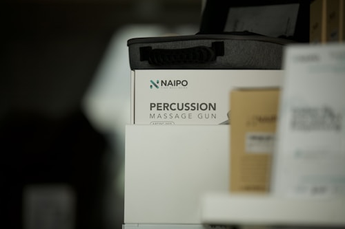 A product display featuring a percussion massage gun in its packaging. The box is predominantly white with the brand and product name clearly visible. There are other boxes partially visible around it, one of which is brown.