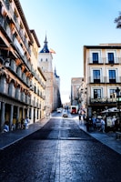 A picturesque street view in a historic European city with traditional buildings lining both sides. The street has cobblestone paving leading up to a prominent building with a pointed tower and the Spanish flag flying atop. People are walking along the street, and cars are driving up the road. The architecture features wrought-iron balconies and varying architectural details, and a vibrant, sunny day illuminates the scene.