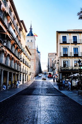 A picturesque street view in a historic European city with traditional buildings lining both sides. The street has cobblestone paving leading up to a prominent building with a pointed tower and the Spanish flag flying atop. People are walking along the street, and cars are driving up the road. The architecture features wrought-iron balconies and varying architectural details, and a vibrant, sunny day illuminates the scene.