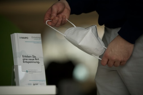 A hand holding a white face mask near a stand displaying brochures. The brochures have some text in German related to relaxation. The person is wearing a dark shirt and light-colored pants.