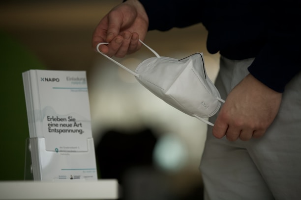 A hand holding a white face mask near a stand displaying brochures. The brochures have some text in German related to relaxation. The person is wearing a dark shirt and light-colored pants.