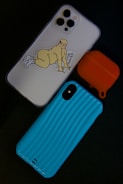 A smartphone with a white case featuring a humorous sticker of a cartoon character in a playful pose is placed alongside an orange AirPods case and a blue phone case with a textured pattern. The arrangement is set against a dark background.