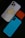 A smartphone with a white case featuring a humorous sticker of a cartoon character in a playful pose is placed alongside an orange AirPods case and a blue phone case with a textured pattern. The arrangement is set against a dark background.