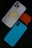 A smartphone with a white case featuring a humorous sticker of a cartoon character in a playful pose is placed alongside an orange AirPods case and a blue phone case with a textured pattern. The arrangement is set against a dark background.