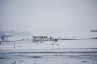 A snowplow is clearing a snow-covered road in a rural area. The landscape is mostly white with snow blanketing the ground and trees. The sky is overcast, creating a cold and wintry atmosphere.
