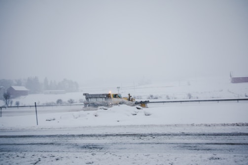 A snowplow is clearing a snow-covered road in a rural area. The landscape is mostly white with snow blanketing the ground and trees. The sky is overcast, creating a cold and wintry atmosphere.