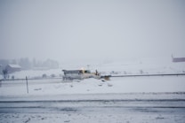 A snowplow is clearing a snow-covered road in a rural area. The landscape is mostly white with snow blanketing the ground and trees. The sky is overcast, creating a cold and wintry atmosphere.
