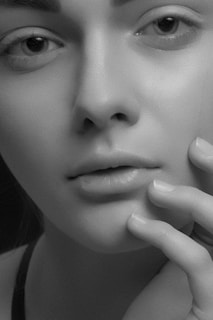A close-up black and white portrait of a person's face showcasing smooth skin, subtle features, and delicate details. The eyes are expressive, and a hand gently touches the face, emphasizing the texture and softness of the skin.