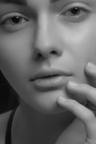 A close-up black and white portrait of a person's face showcasing smooth skin, subtle features, and delicate details. The eyes are expressive, and a hand gently touches the face, emphasizing the texture and softness of the skin.