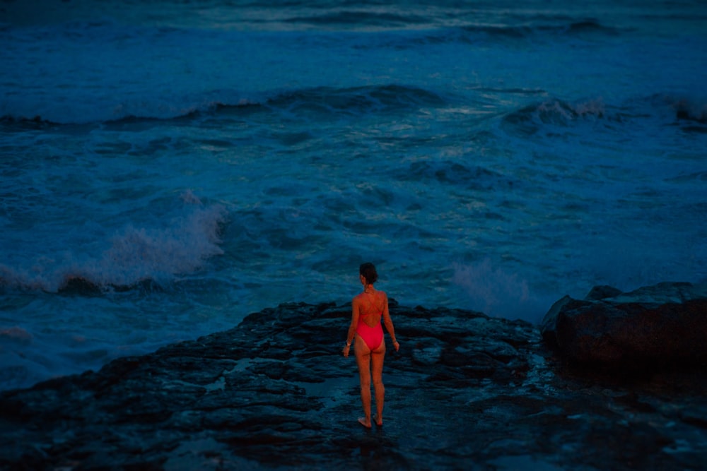 woman in red dress standing on rock near ocean waves during daytime