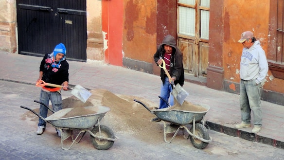 Three people are working outdoors with wheelbarrows filled with sand, appearing to be involved in a construction or repair task. One person is shoveling sand, another is holding a shovel, and the third is standing by observing. The backdrop includes a rustic building with worn paint, featuring a dark door and a pair of light wooden doors.