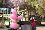 A vibrant street market scene featuring a cotton candy stand with bags of pink cotton candy hanging. A vendor in a red cap stands behind the stand. The background includes a stone fountain with spouting water and lush trees with purple and green foliage. The market area is bustling with pedestrians.
