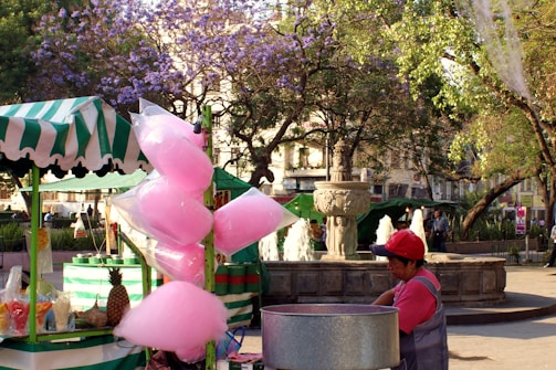 A vibrant street market scene featuring a cotton candy stand with bags of pink cotton candy hanging. A vendor in a red cap stands behind the stand. The background includes a stone fountain with spouting water and lush trees with purple and green foliage. The market area is bustling with pedestrians.