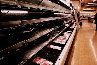 An aisle in a grocery store with a nearly empty refrigerated display case. A few packages of meat remain on the shelves. The scene is dimly lit, creating a stark contrast with the brightly lit shelves. A single shopper is visible in the distance.