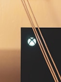 A close-up of a gaming console showing the illuminated power button with the signature logo. Three braided cables are hanging across the front, contrasting against the sleek black surface of the console.