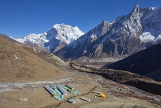 A mountainous landscape with snow-capped peaks under a clear blue sky. In the foreground, a small settlement with colorful tents and simple shelter structures sits amidst the rocky terrain. The area surrounding the settlement consists of dry, brown earth, contrasting with the majestic, rugged mountains that stretch into the distance.