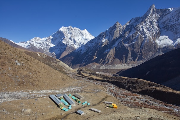 A mountainous landscape with snow-capped peaks under a clear blue sky. In the foreground, a small settlement with colorful tents and simple shelter structures sits amidst the rocky terrain. The area surrounding the settlement consists of dry, brown earth, contrasting with the majestic, rugged mountains that stretch into the distance.