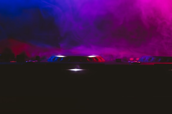 The image features a DJ turntable or mixer on a dark stage, enveloped in a colorful mist. The lighting creates a dramatic effect with blue and pink hues swirling around the equipment.