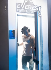 A person wearing headphones and gloves stands in the doorway of a cryotherapy chamber, surrounded by mist. The chamber is branded with the name 'Everest Powered by Cryobuilt'. The individual is dressed in a sleeveless top and athletic pants, and has visible tattoos on one arm.