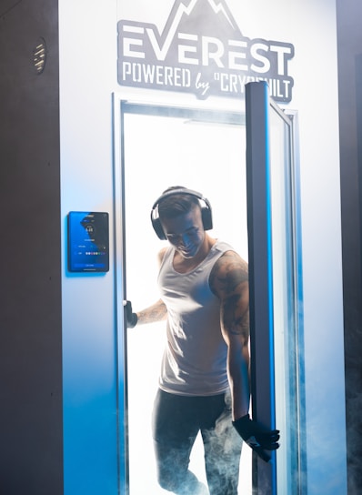 A person wearing headphones and gloves stands in the doorway of a cryotherapy chamber, surrounded by mist. The chamber is branded with the name 'Everest Powered by Cryobuilt'. The individual is dressed in a sleeveless top and athletic pants, and has visible tattoos on one arm.