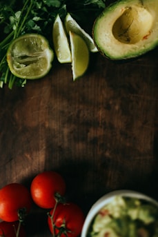 A wooden surface features an array of fresh ingredients including sliced avocado, lime wedges, tomatoes on the vine, cilantro, and a small bowl of guacamole.