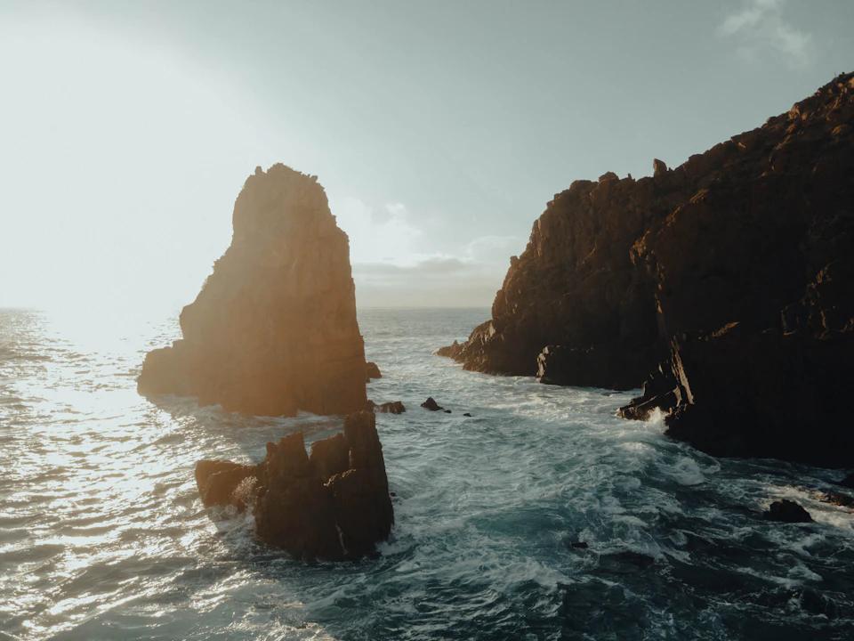 The image depicts a rocky coastline with two large, jagged rocks protruding from the water's surface. The rocks are situated on the left side of the frame and appear to be part of an underwater landscape. The sky above is a light blue color, suggesting a clear day. The ocean below is a darker shade of blue, indicating that it may be deeper or colder than the surface level. The image does not contain any discernible text or human-made objects, making the scene appear untouched and natural.