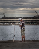 A person stands on a concrete walkway by the water, holding a fishing line near a fishing rod. They are wearing a cap, a brown and white striped shirt, and camouflage shorts. The background features a large, industrial building and a crane under a cloudy sky.