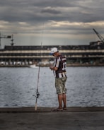 A person stands on a concrete walkway by the water, holding a fishing line near a fishing rod. They are wearing a cap, a brown and white striped shirt, and camouflage shorts. The background features a large, industrial building and a crane under a cloudy sky.