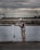 A person stands on a concrete walkway by the water, holding a fishing line near a fishing rod. They are wearing a cap, a brown and white striped shirt, and camouflage shorts. The background features a large, industrial building and a crane under a cloudy sky.